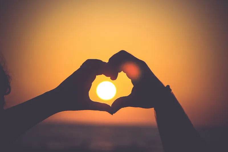 Two hands from two different people form a heart around a sunset in the background
