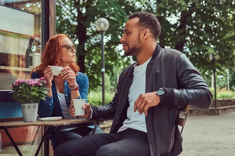 Two people sit at a table drinking coffee outside