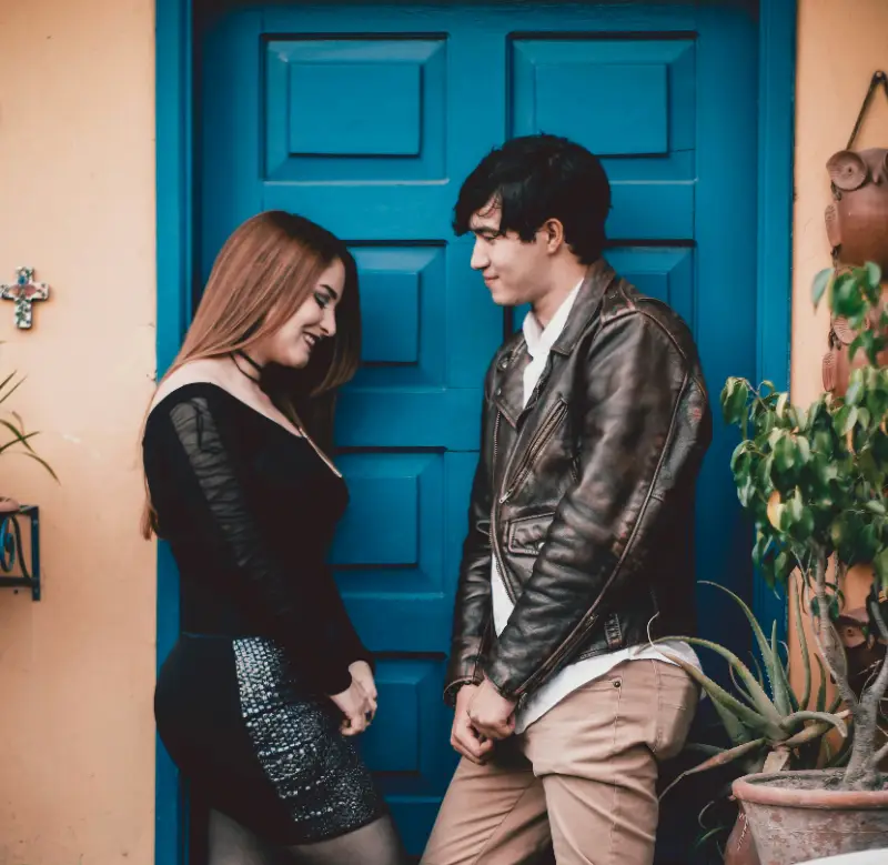 A man and a woman stand in front of a door, looking at each other and sharing a moment