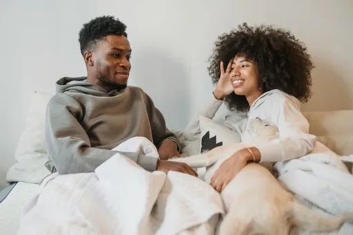 A man and a woman in bed, fully-clothed and talking. The man looks bemused, and the woman is smiling at him.