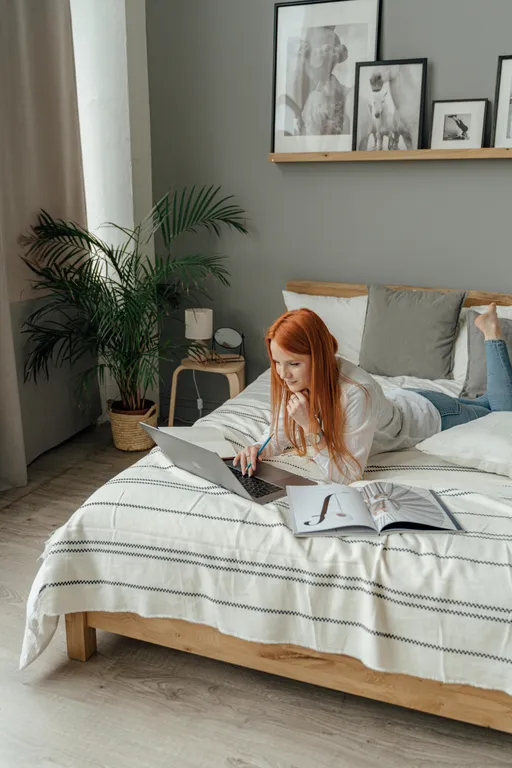 An orange-haired woman laying bed working on a laptop in a brightly lit bedroom