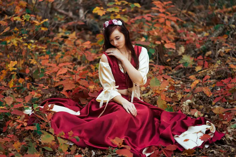 A brunette woman in a red and white dress sits pensively on a bed of autumn leaves
