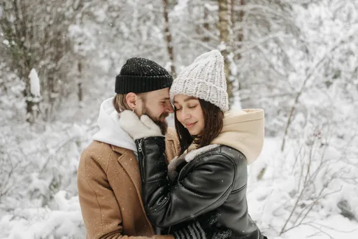A loving couple holding each other in wintery woods, with snow falling all around them. They are bundled up and look very happy.