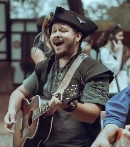 A bard plays the guitar in a Ren Faire while people in medieval garb pass by.