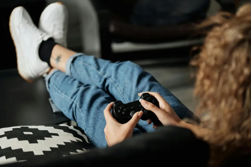A woman is sitting on the couch with a PS2 controller in a relaxed pose, playing video games.